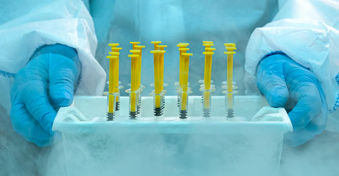 The true cost of cold storage failures - Scientist wearing protective gloves and lab suit holding frozen medical syringes in a cold storage container with visible frost and vapor.